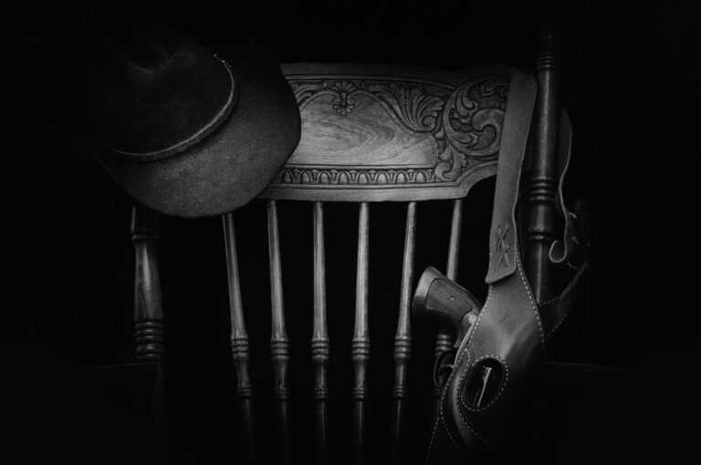 A classic western-themed black and white still life featuring a hat, wooden chair, and gun.