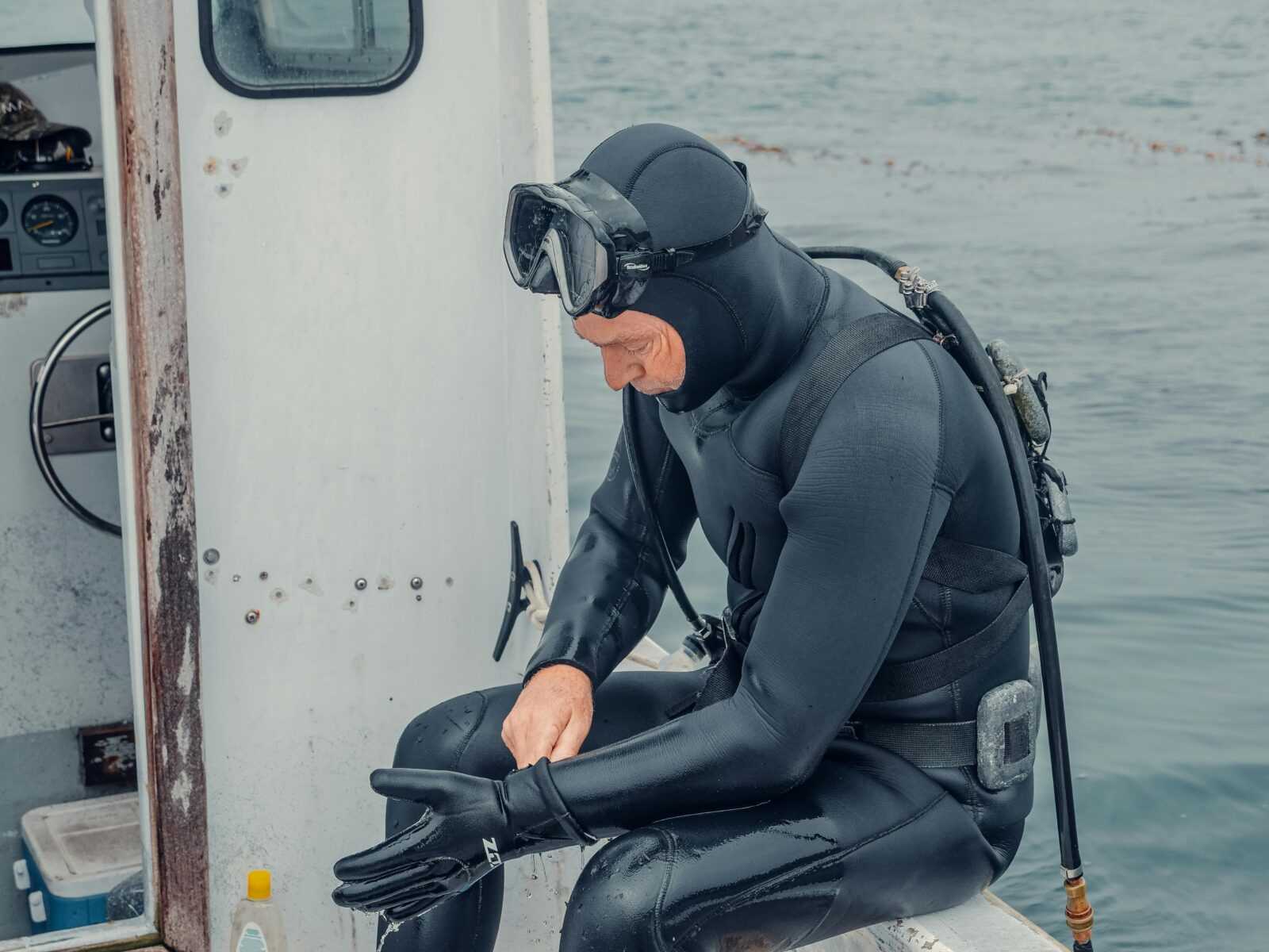 A diver putting on gloves while sitting on the edge of a boat near the water