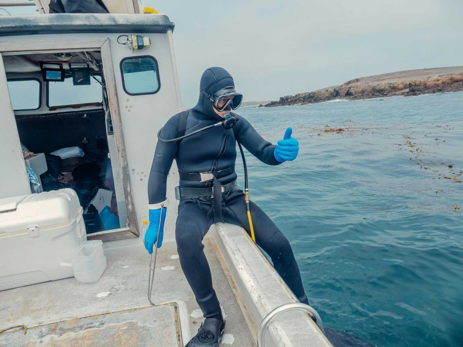 A diver with a wetsuit giving a thumbs up while sitting on the edge of a boat near the water